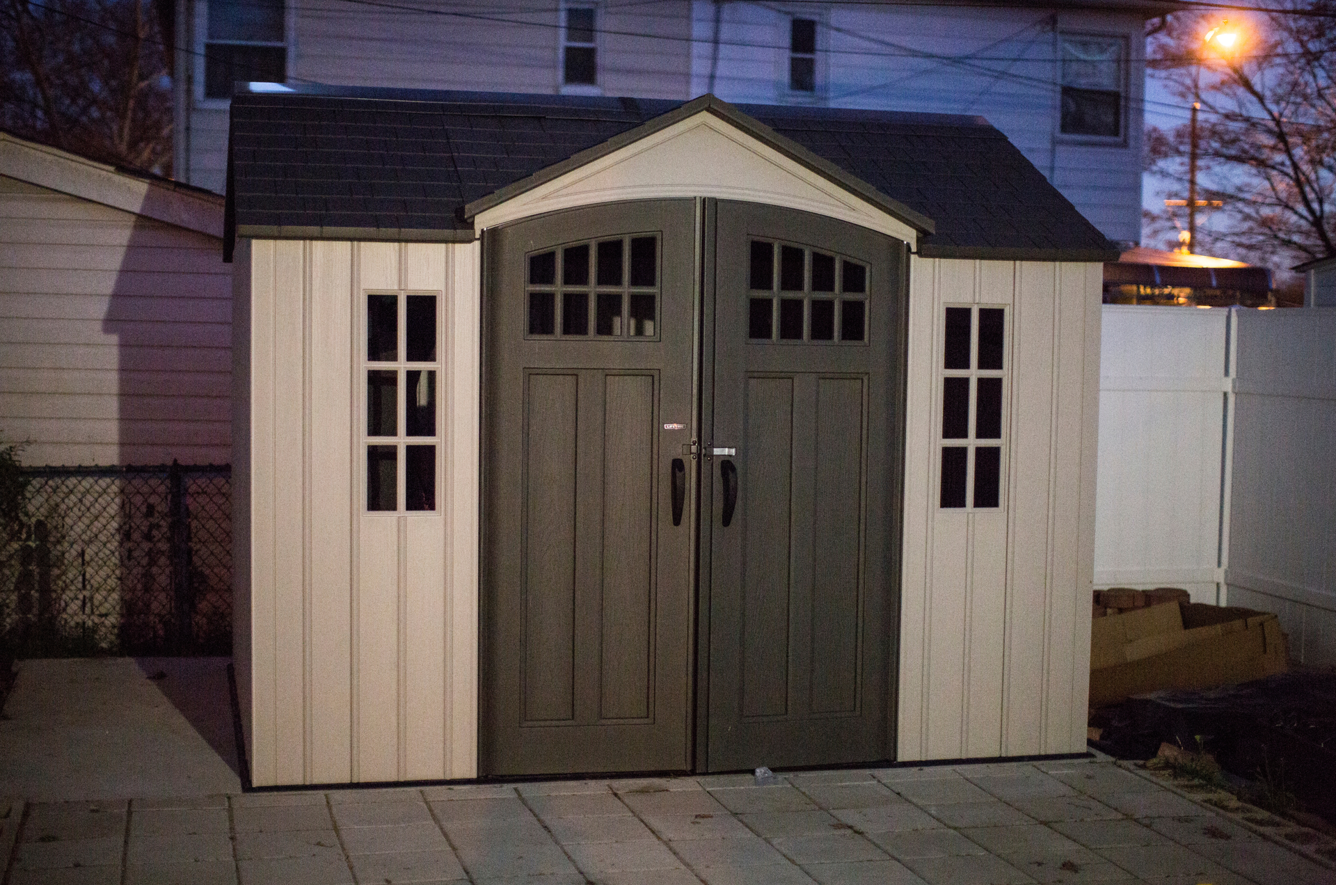Tan and gray shed with double doors and two small windows at dusk.