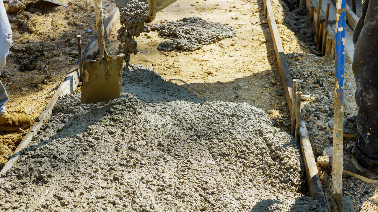 Wet concrete is poured into a wooden frame at a construction site, with a shovel standing nearby.