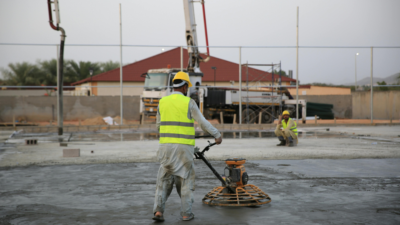 A worker uses a concrete power trowel on a construction site, while a second person sits in the background.