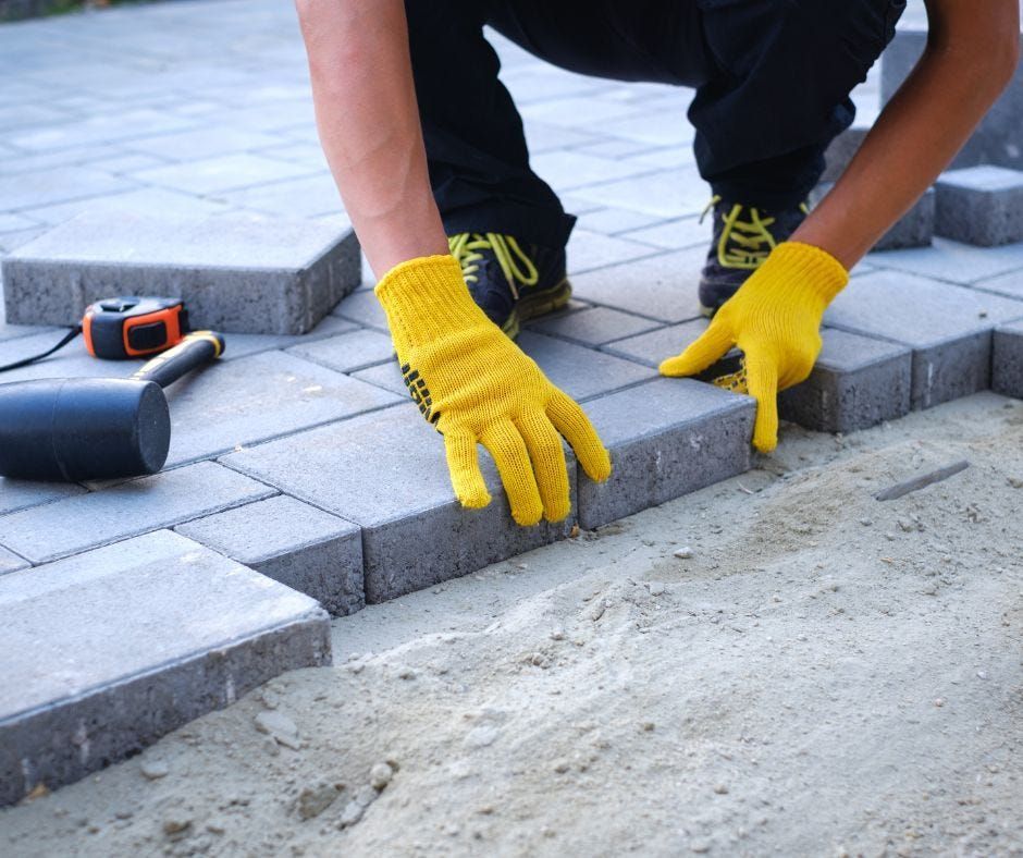 A person wearing yellow gloves installs grey paving stones on a sandy base, with a rubber mallet and tape measure nearby.