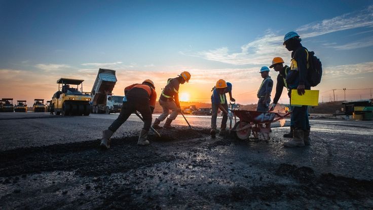A group of construction workers in hard hats and safety gear working on a road surface at sunset with machinery nearby.