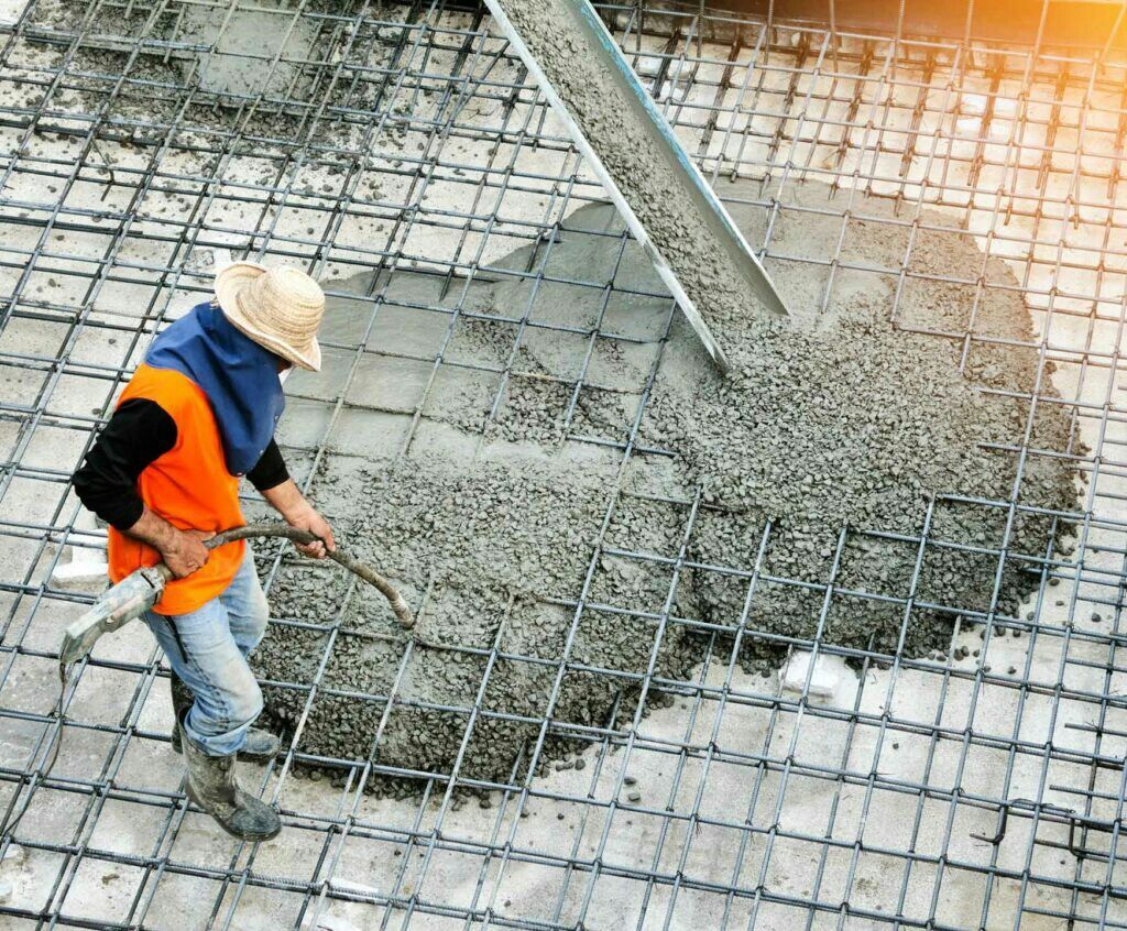A construction worker in a hard hat and safety vest directs wet concrete poured from a chute onto a rebar grid.