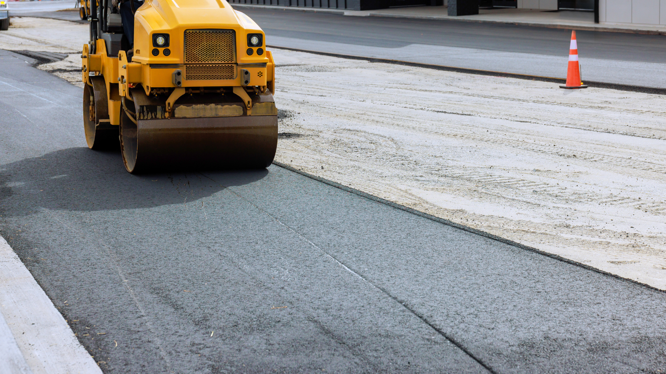 A yellow road roller compacts freshly laid asphalt on a construction site next to an orange traffic cone.
