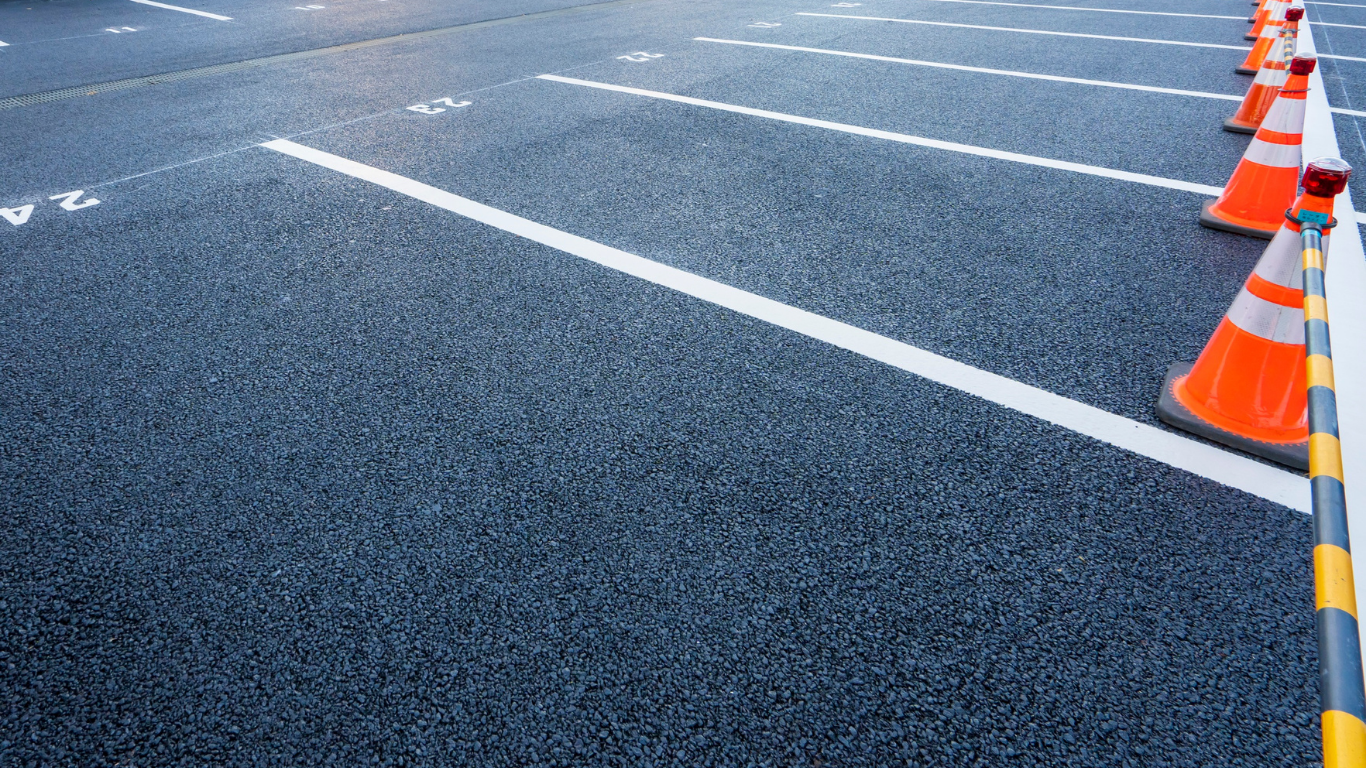 A series of orange traffic cones connected by a striped pole lines a white parking space marking on gray asphalt.