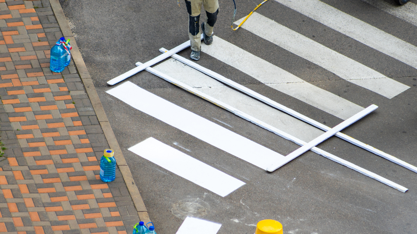 A person repainting a white crosswalk stripe on an asphalt road using a metal stencil frame.