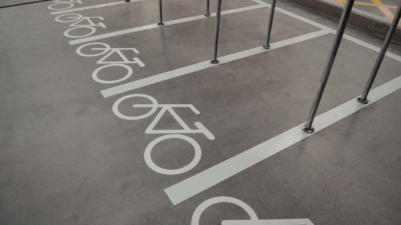 Bicycle parking spots marked with white icons on a gray concrete surface, featuring metal racks for securing bikes.