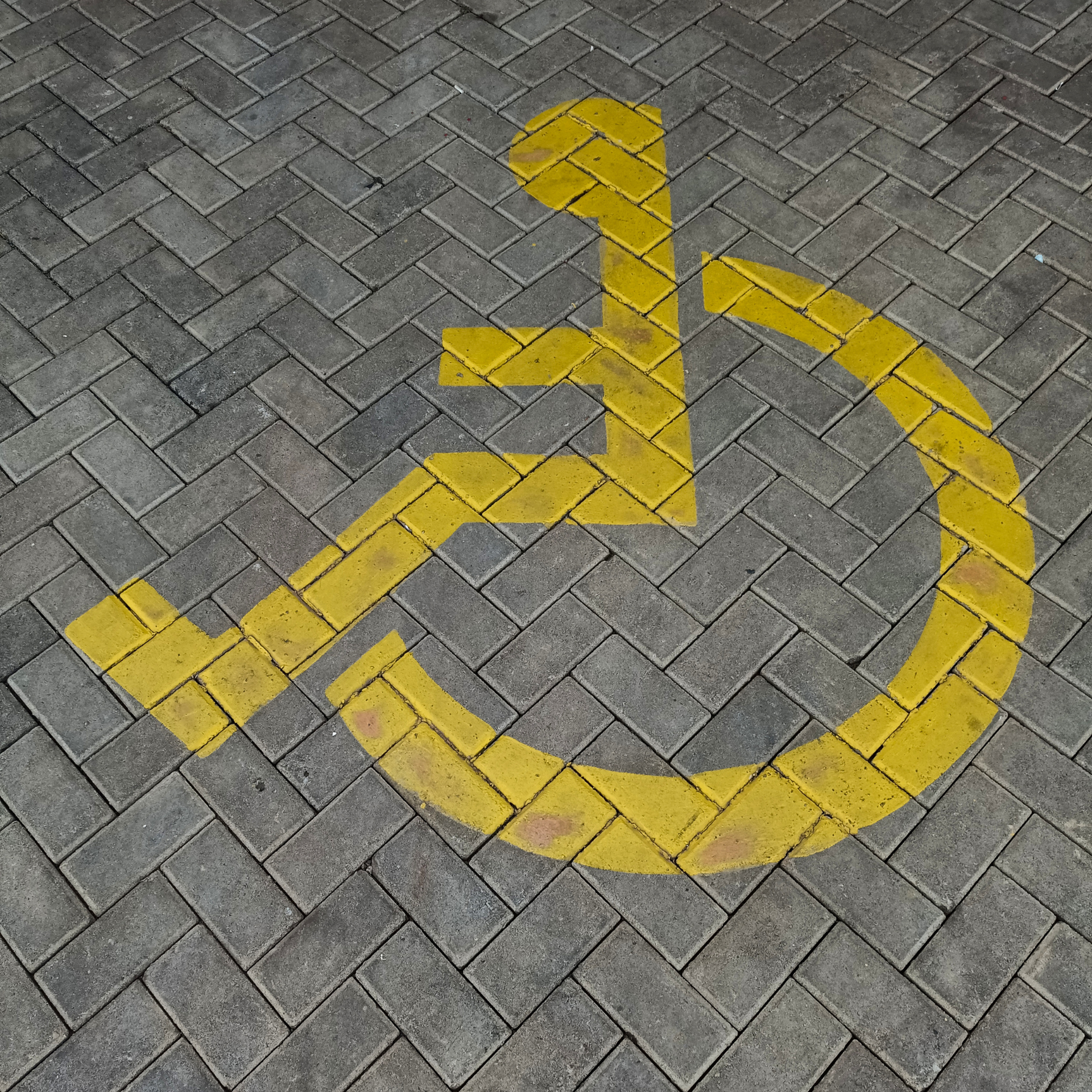 Yellow wheelchair accessibility symbol painted on grey herringbone-patterned brick pavement.