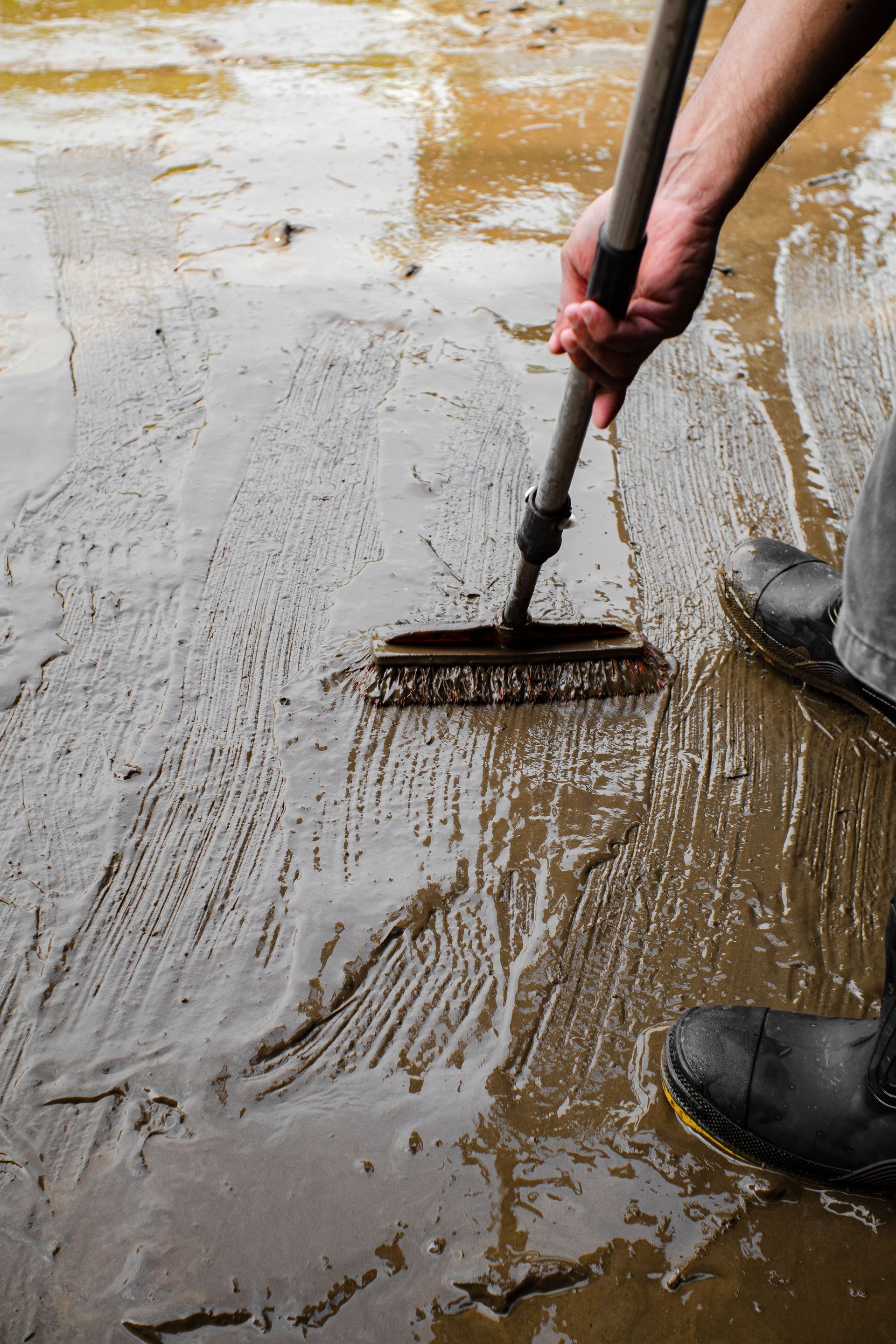 Person raking wet mud across a flooded surface with a metal rake and boots visible