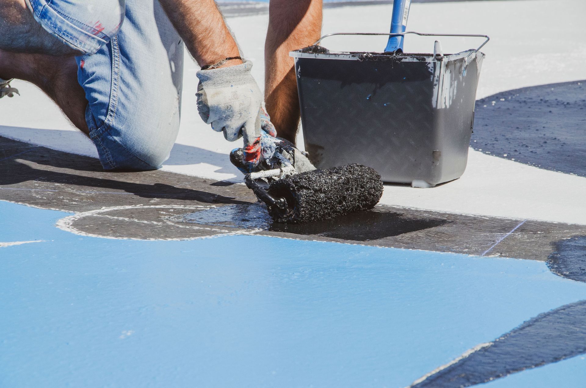 Person applying black coating with a roller on a blue roof, beside a paint bucket