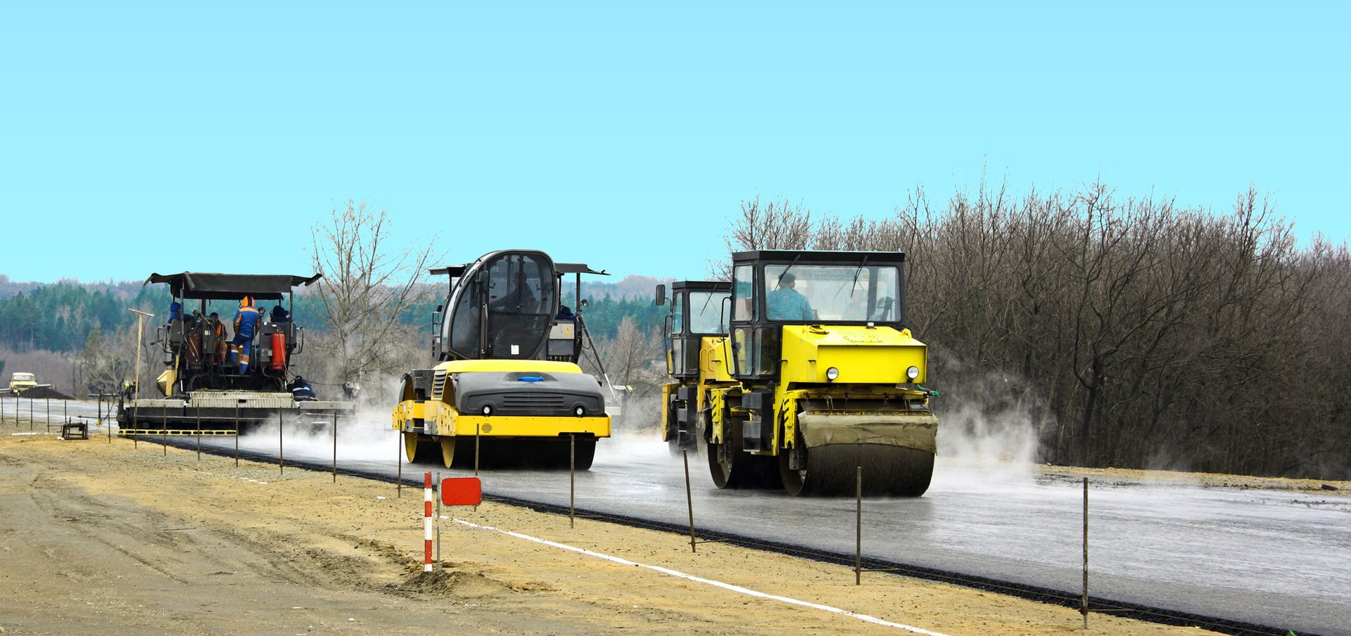 Three heavy construction machines paving a new road on a clear day, with steam rising from the fresh asphalt.