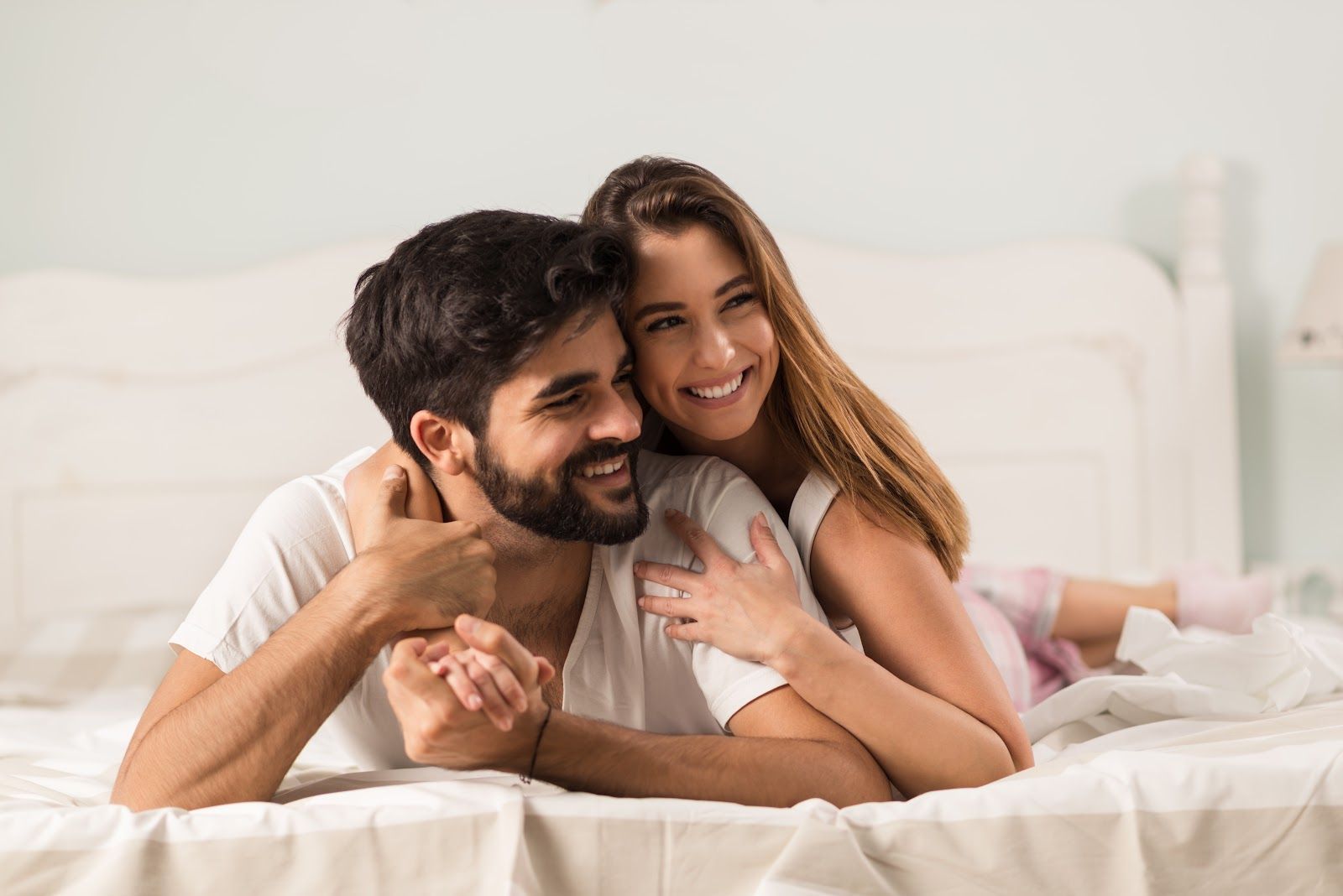 Couple embracing and smiling on a bed, woman's arms around man, looking happy.