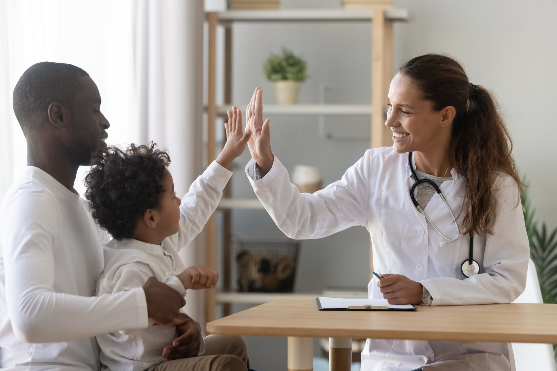 A doctor gives a high-five to a young patient sitting on a parent's lap in a bright office.
