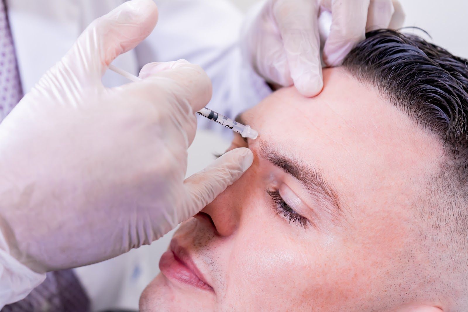 Person receiving cosmetic injections in the forehead; gloved hand administers treatment with a syringe.