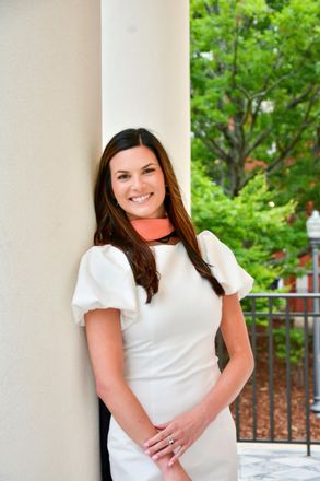 Woman in white dress smiles, leaning against a pillar. She has long brown hair and an orange scarf. Outdoors.