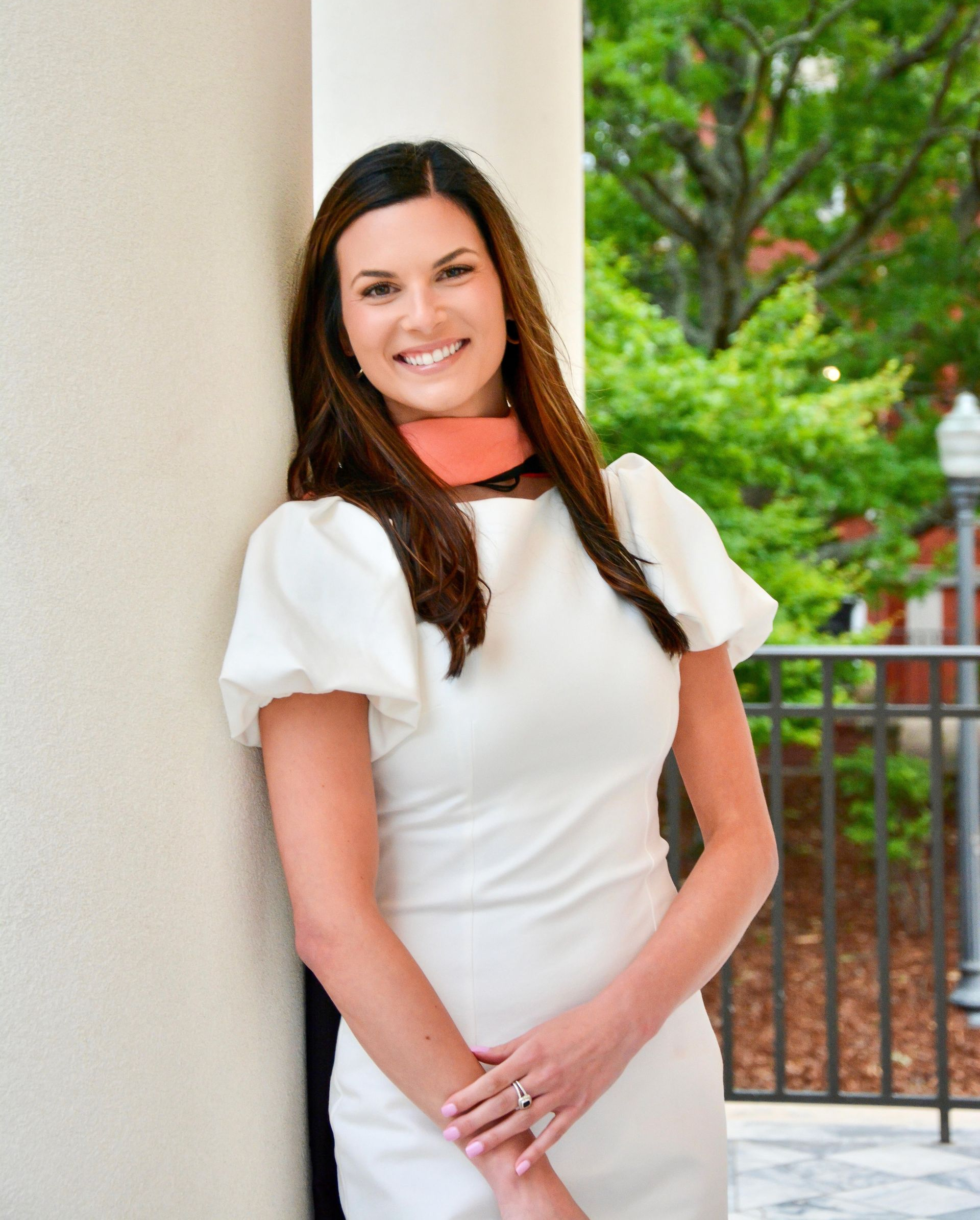Woman in white dress smiles, leaning against a pillar. She has long brown hair and an orange scarf. Outdoors.