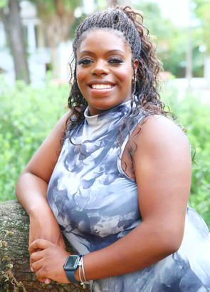 Smiling Black woman with curly hair, leaning on a tree branch outdoors, wearing a patterned sleeveless top.