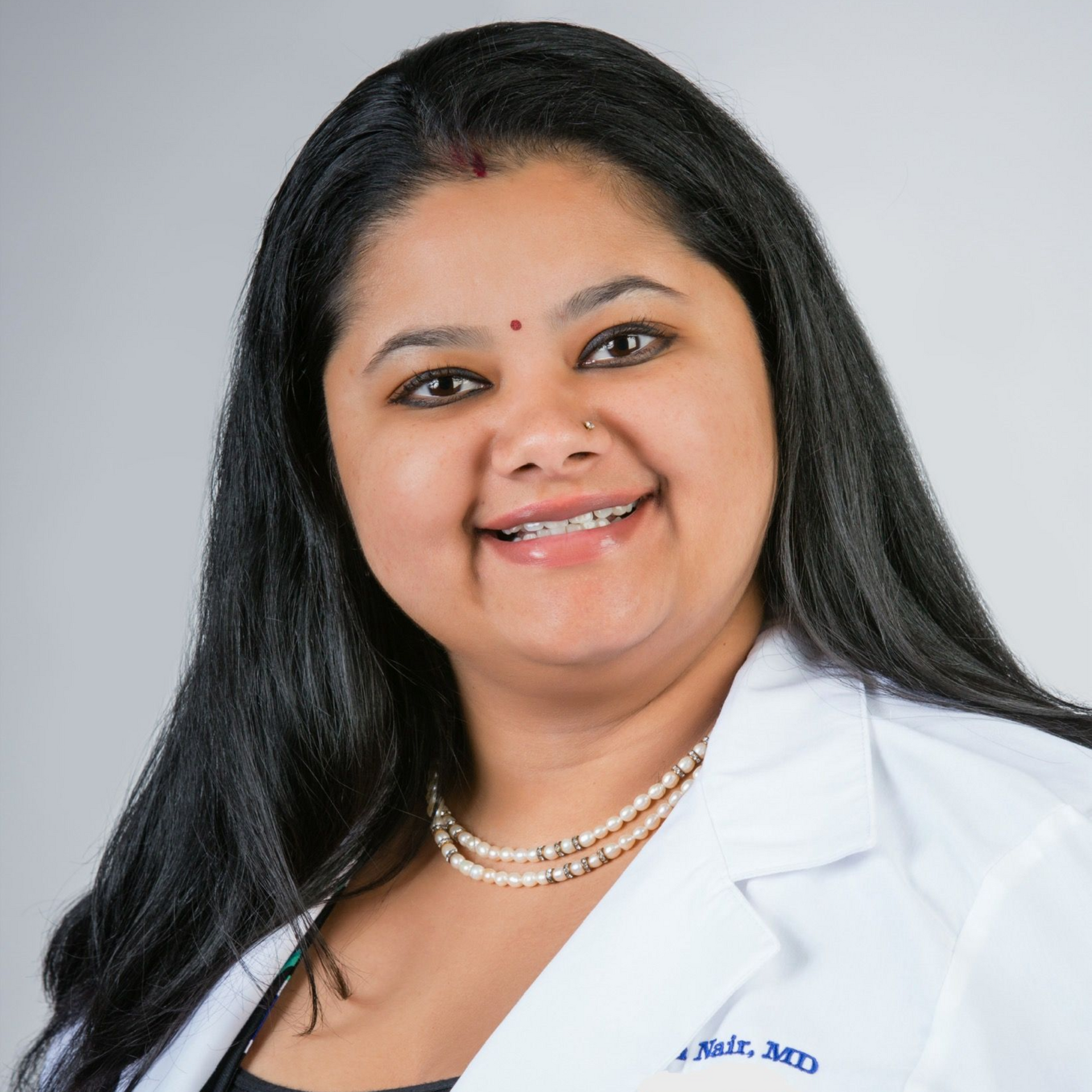 Woman in white lab coat smiles; pearl necklace.
