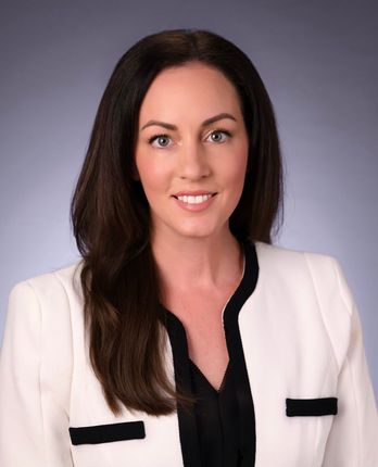 Woman with brown hair, in a white blazer with black trim, smiling.