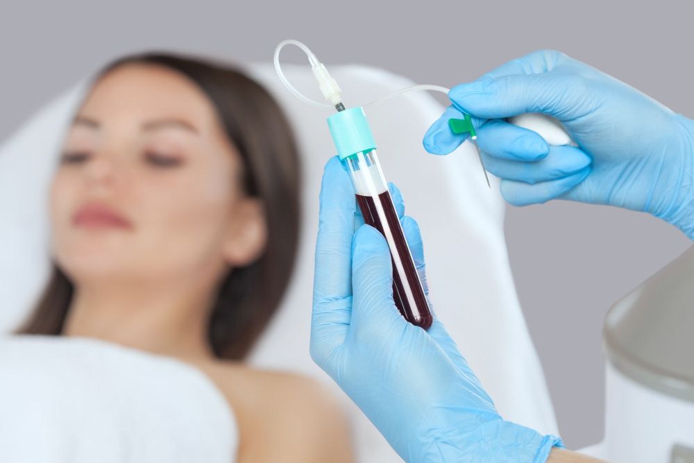 Person receiving blood draw. Hands in blue gloves holding tube of blood and needle. Light setting.