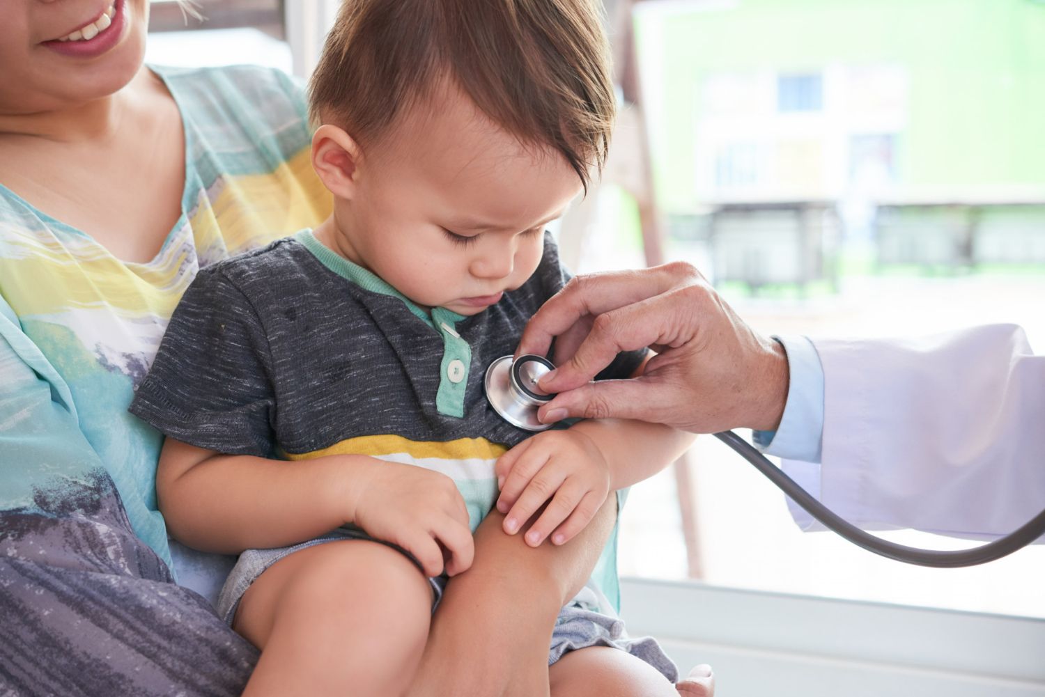 Doctor listening to baby's chest with a stethoscope, mother holding baby, in a medical setting.