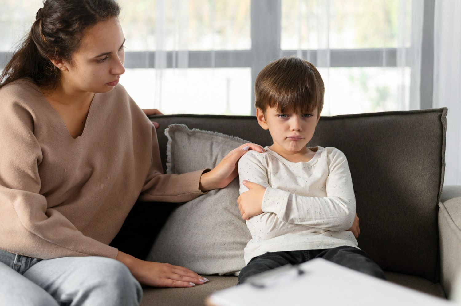 Woman comforts a young, frowning boy on a couch. Setting is indoors, with papers visible.