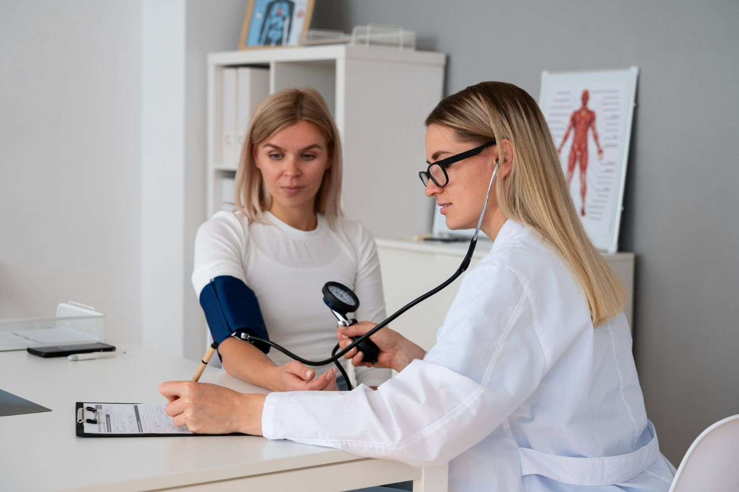 Doctor taking patient's blood pressure in an office. Doctor is blonde, wearing glasses and a white coat.