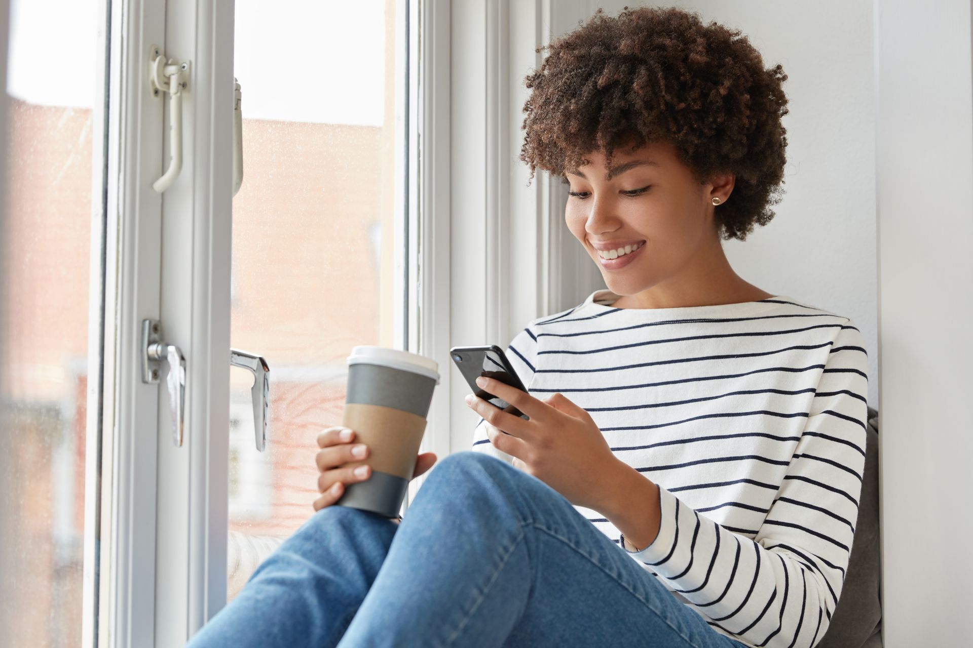 A woman is sitting on a window sill holding a cup of coffee and a cell phone.