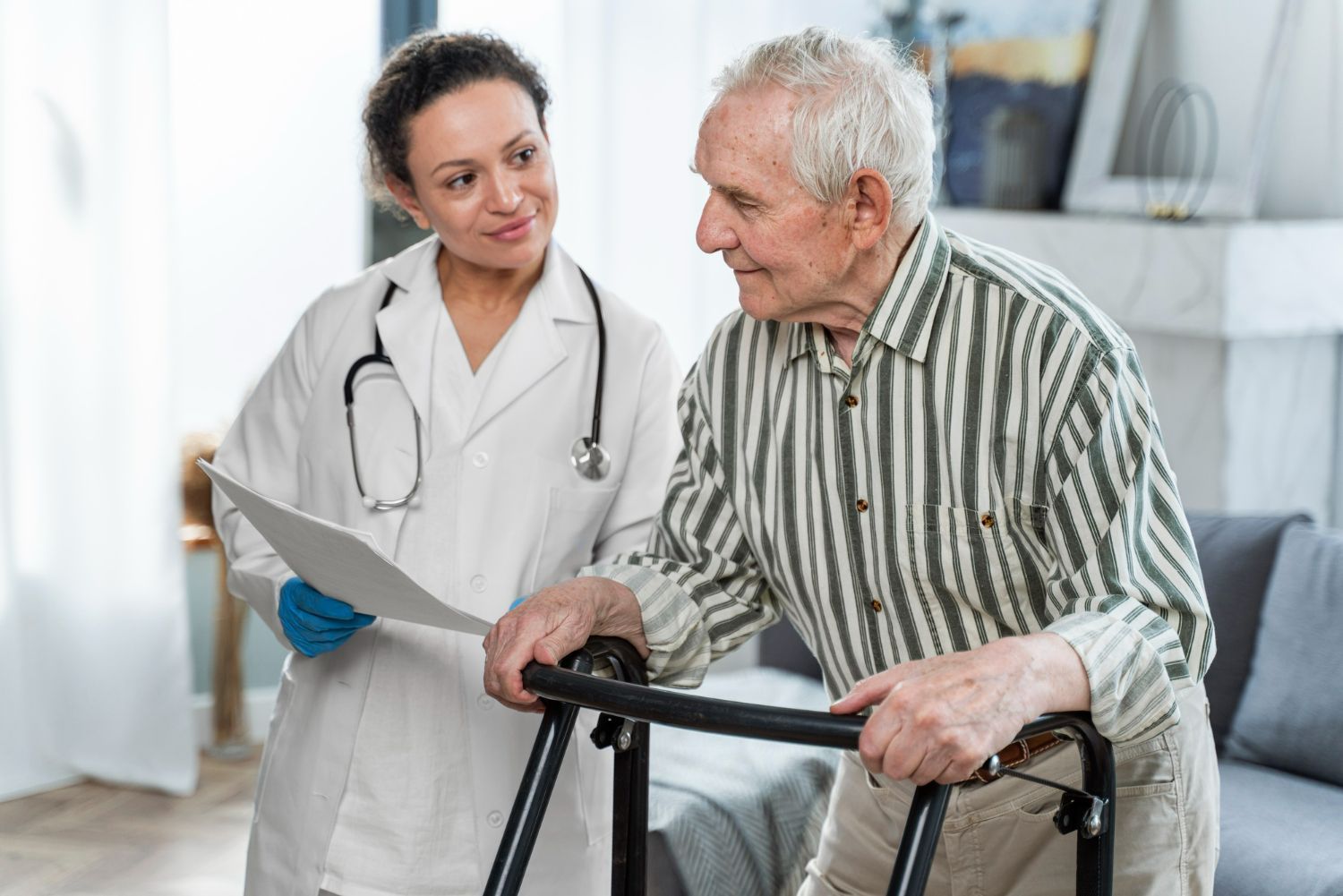 A doctor in a white coat assists an elderly man with a walker in a living room.