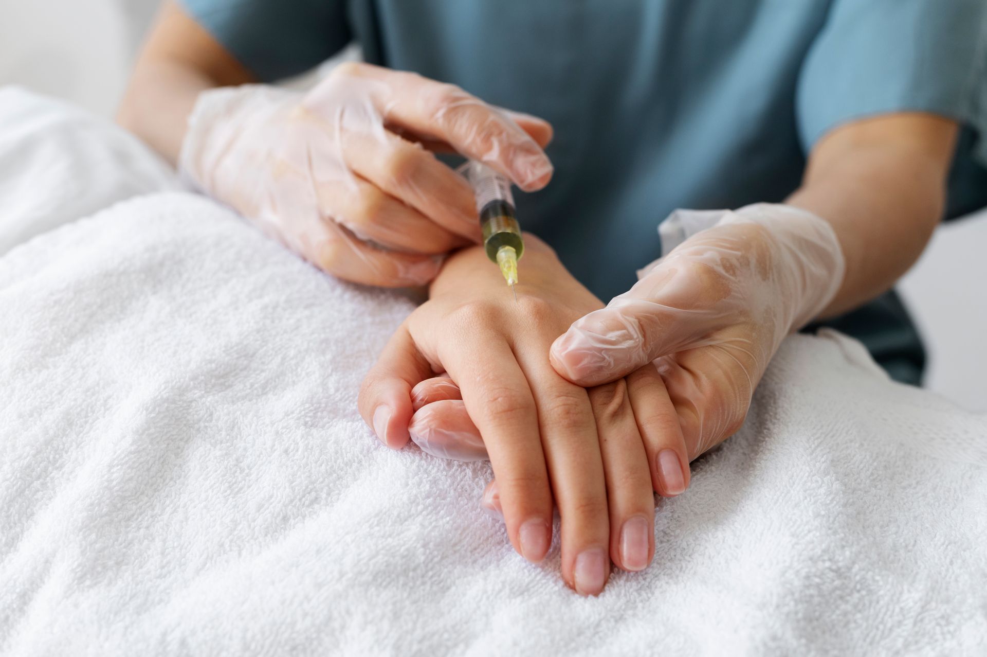 Gloved hands administering an injection into the back of a patient's hand, on a white towel.