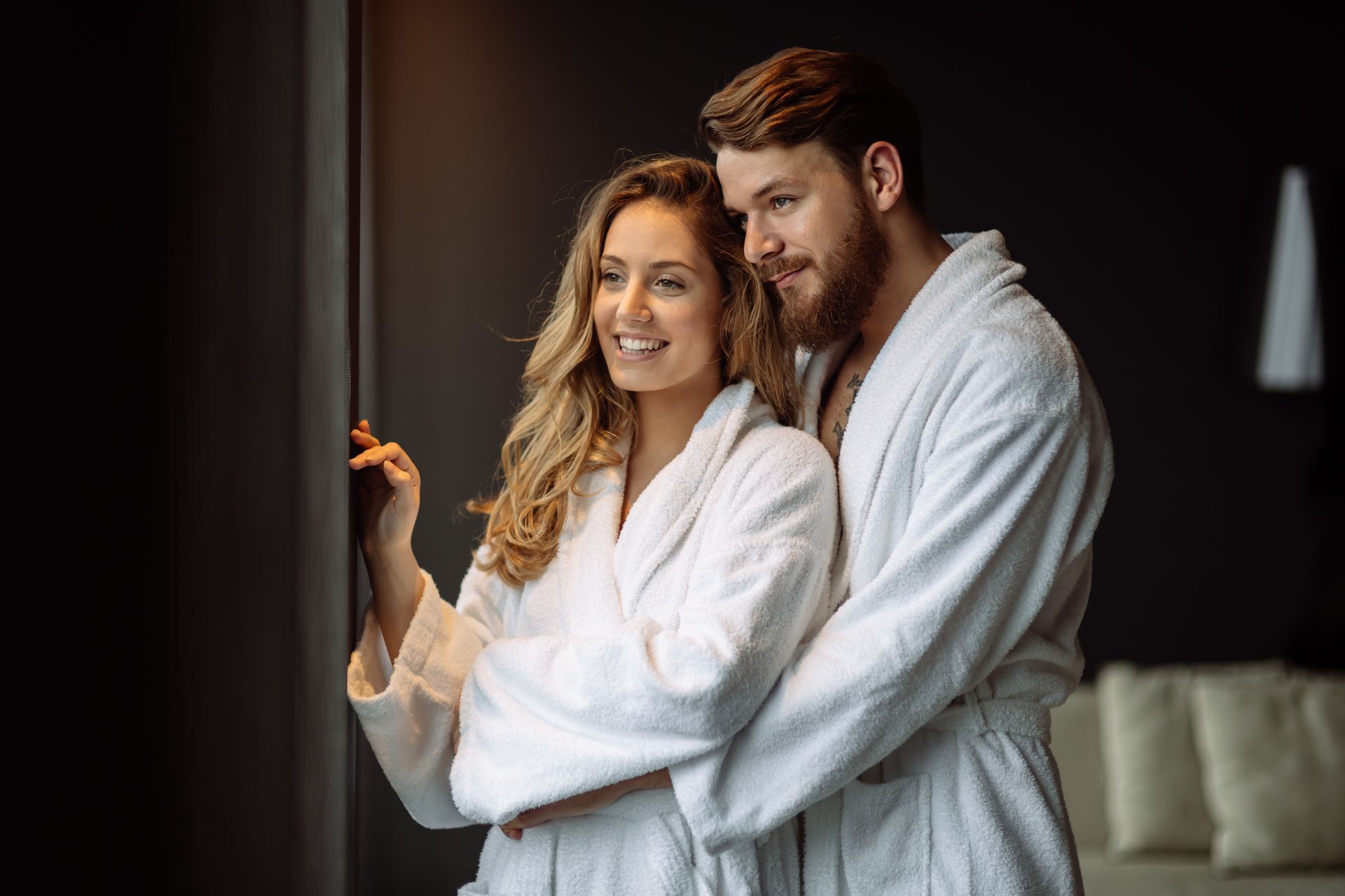 A man and a woman in bathrobes are looking out of a window.