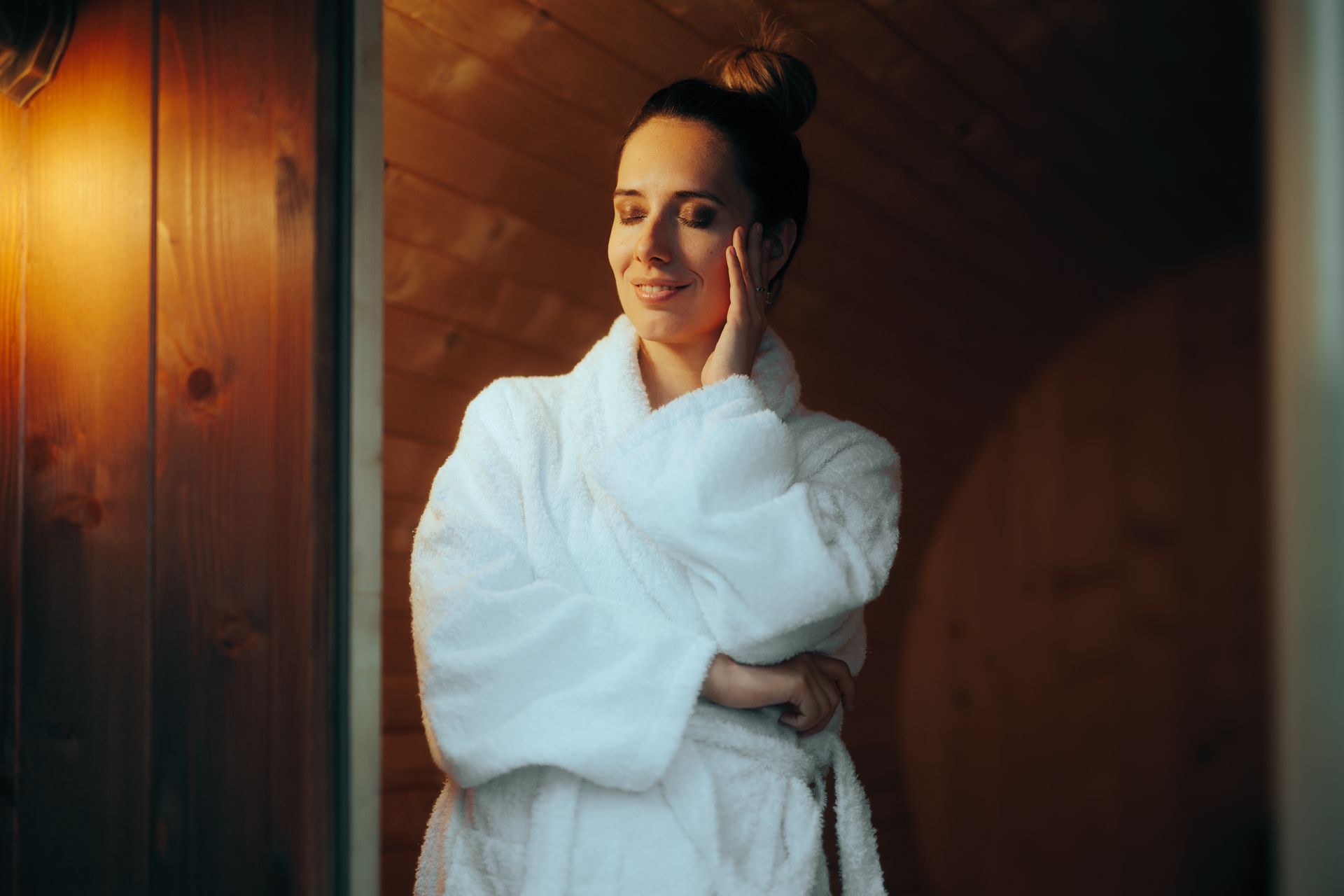 Person in a white robe smiling in a warmly lit wooden room, hand on cheek.
