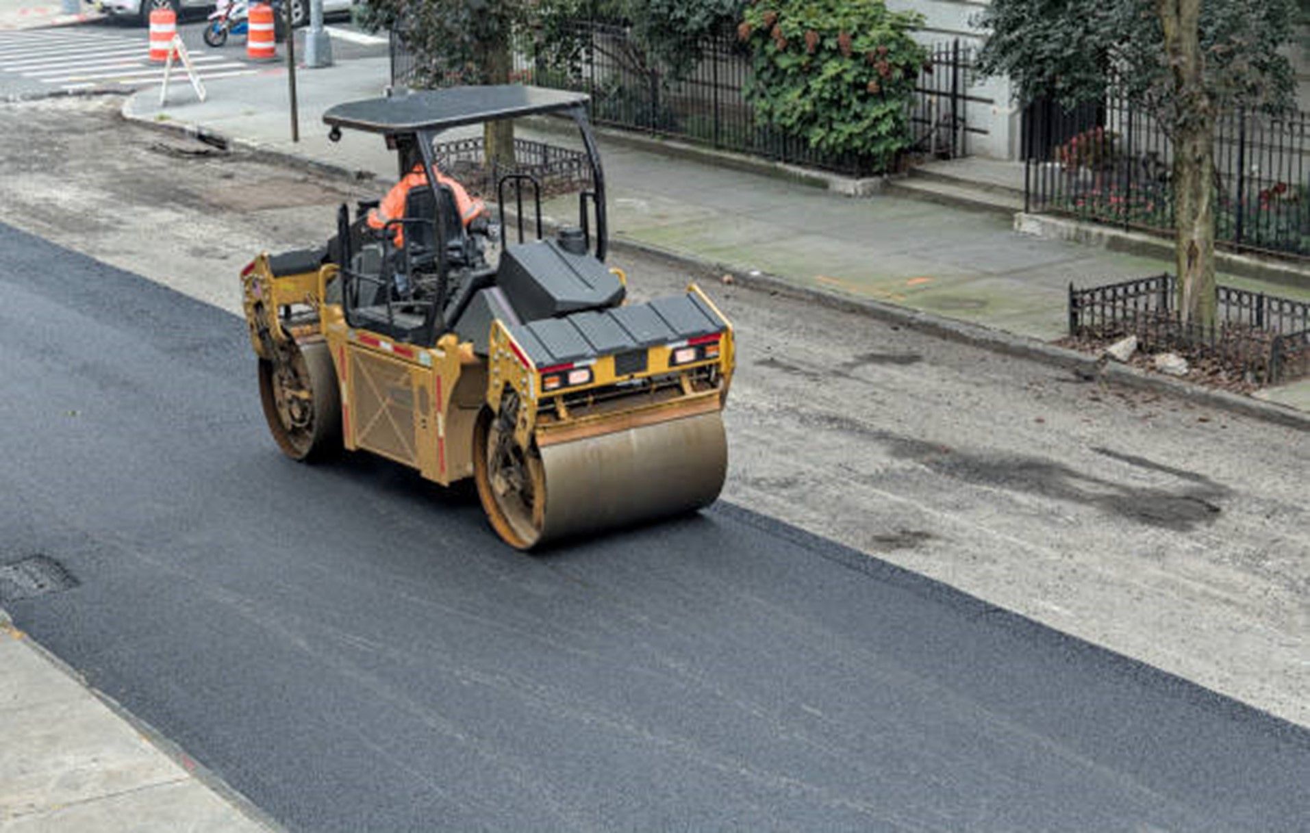 Road roller compacting fresh asphalt on a city street, operated by a worker wearing an orange safety vest.