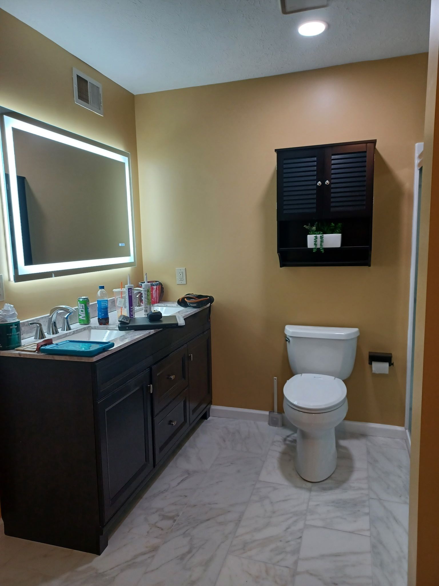 Bathroom with dark vanity, illuminated mirror, black wall cabinet, and white toilet. Light tile floor and tan walls.