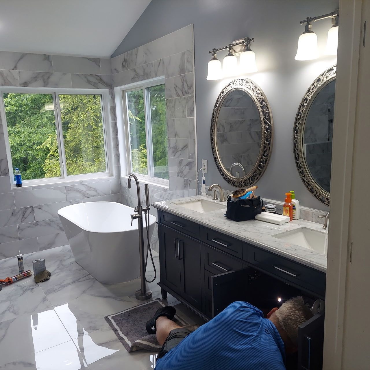 Plumber working on bathroom plumbing; a marble-tiled, modern bathroom. Includes a bathtub, vanity with mirrors, and a window.