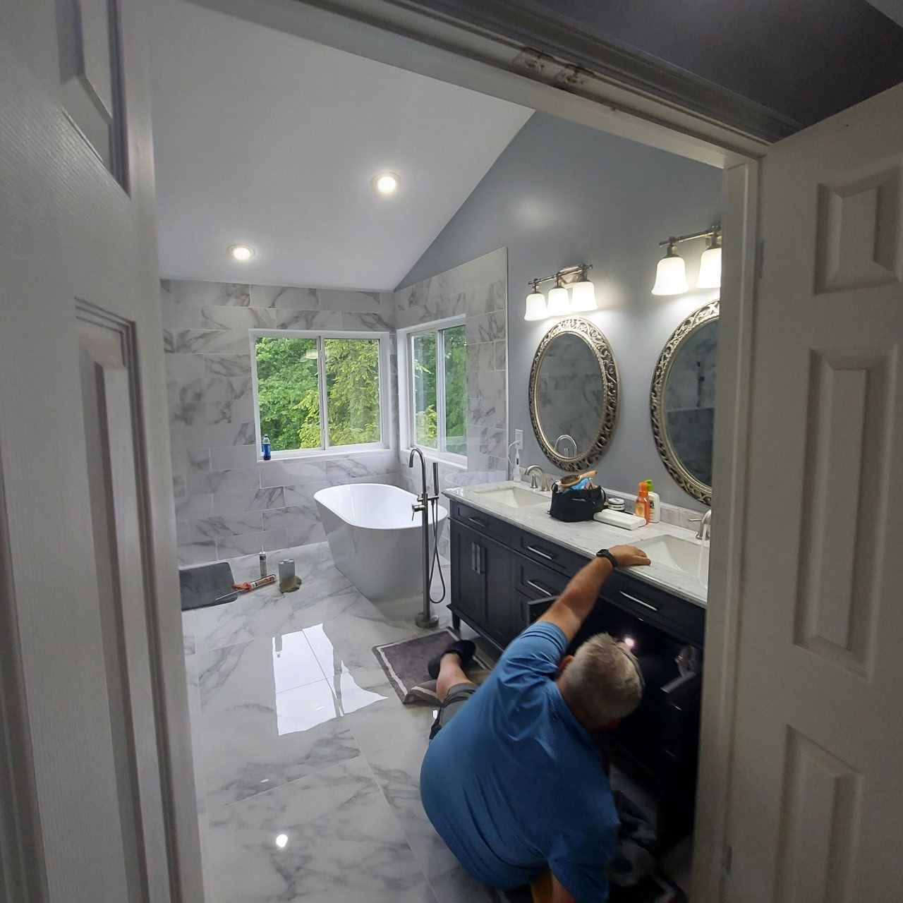 Plumber working under a bathroom vanity. The bathroom has marble tile and a white tub.