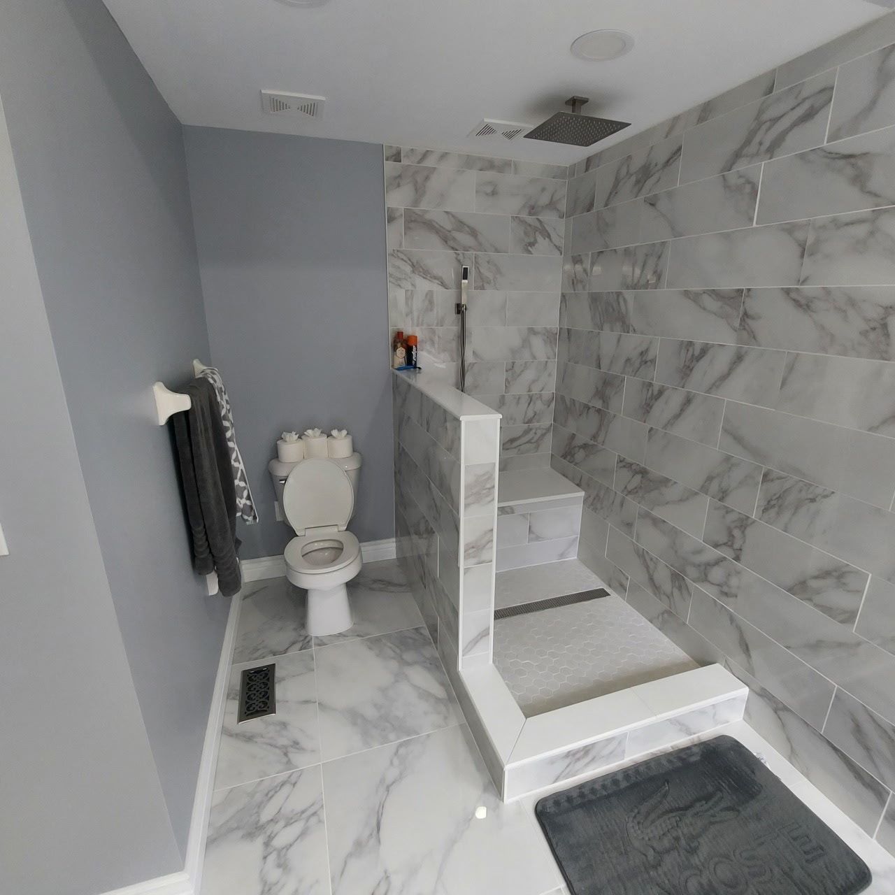 Bathroom with marble-look tile, shower, toilet, and towel rack. Gray walls and a dark gray bath mat.