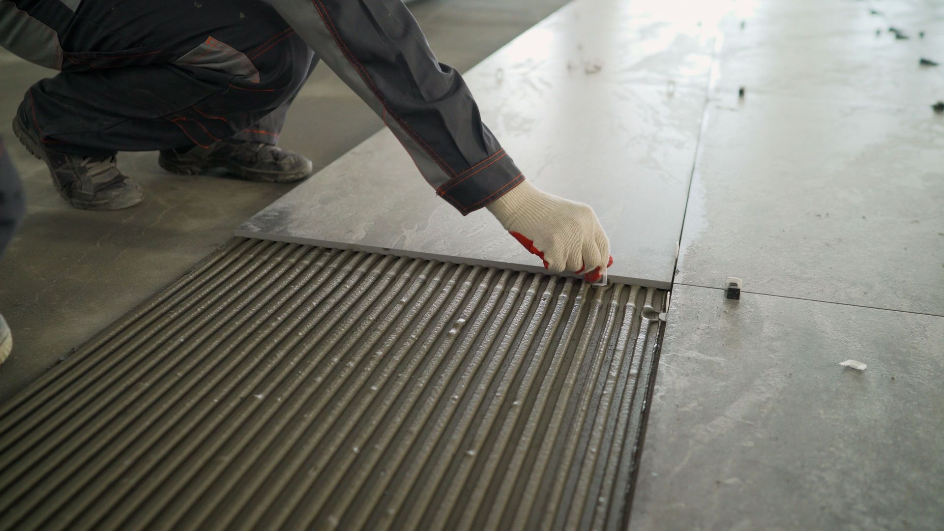 Person applying tile, spreading grout with a notched trowel on a concrete floor.