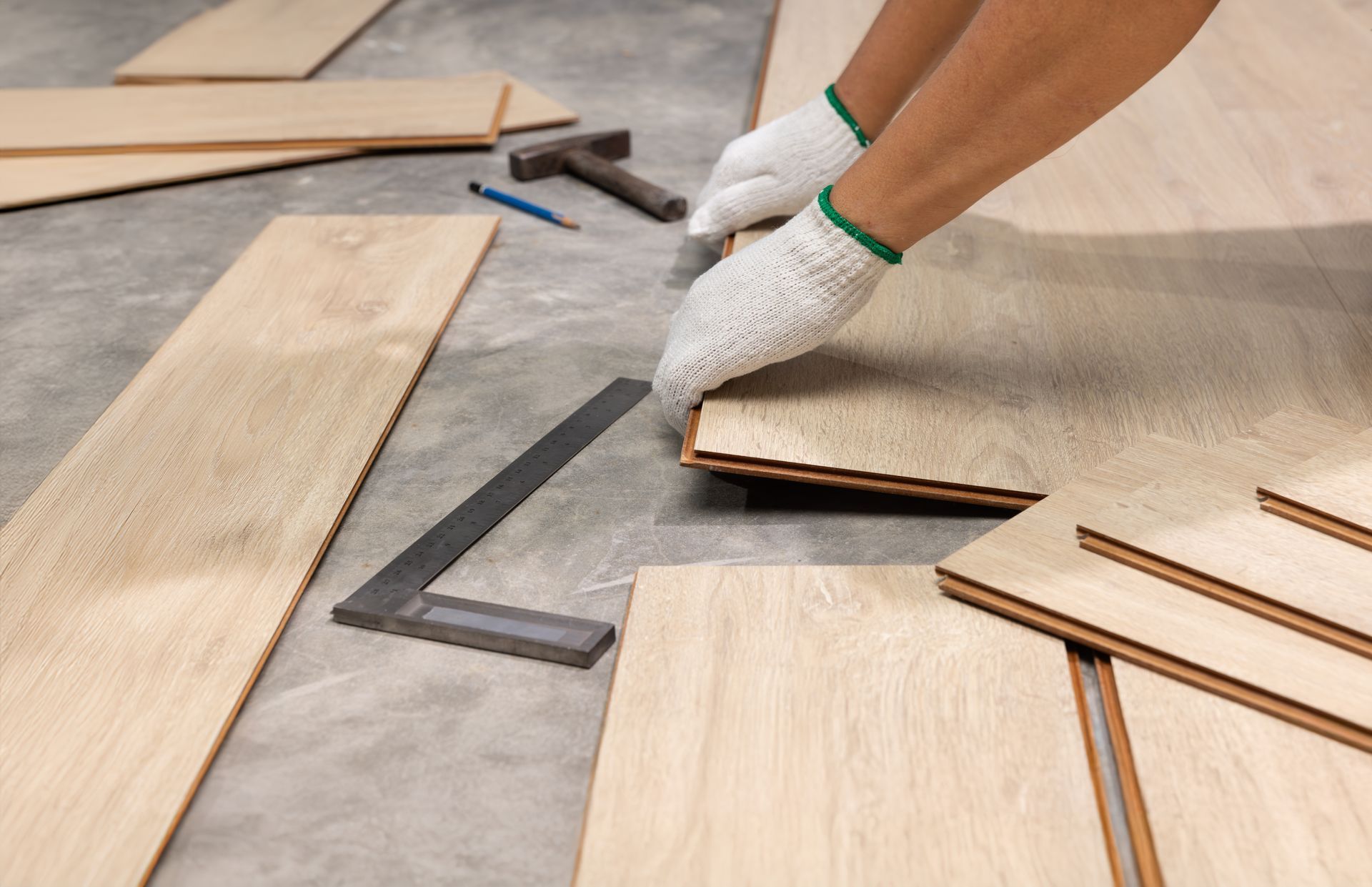 Person wearing gloves, installing wood flooring, using a square, hammer, and pencil.