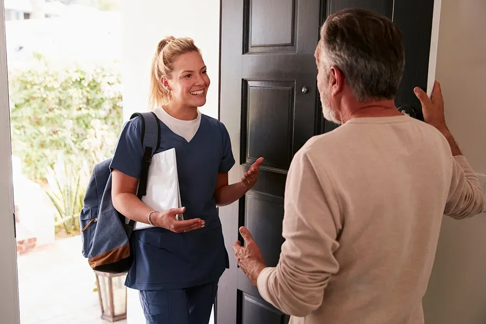 Woman in scrubs with a backpack greets a man at a doorway, smiling and gesturing.