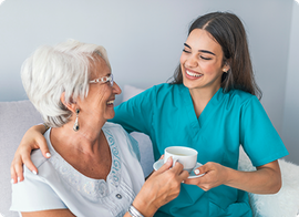 Woman in teal scrubs offers a cup to an elderly woman with short gray hair; both are smiling.