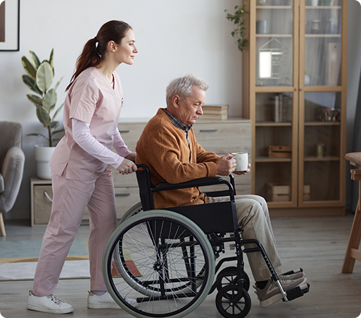 Caregiver pushing a person in a wheelchair indoors, holding a mug. The caregiver wears pink scrubs.