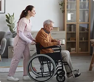 Caregiver pushing a person in a wheelchair indoors; holding a mug, and wearing a brown sweater.