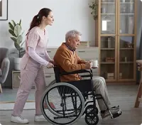 Caregiver pushing a person in a wheelchair indoors; holding a mug, and wearing a brown sweater.