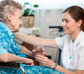 Caregiver offers water to an elderly person in a wheelchair indoors.