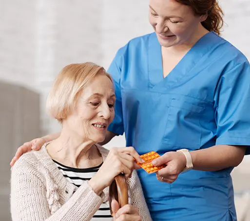 Nurse handing pills to an older adult. Woman in blue scrubs, other in sweater, holding a cane.
