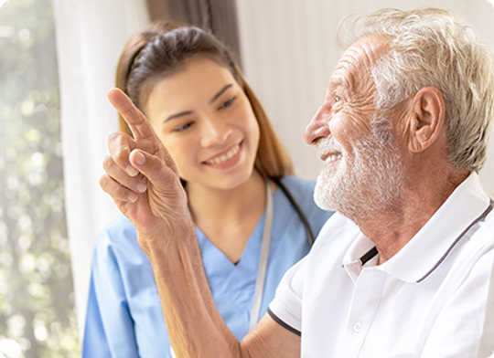 Nurse points, smiling, to something out of frame; elderly man smiles and looks in the same direction.