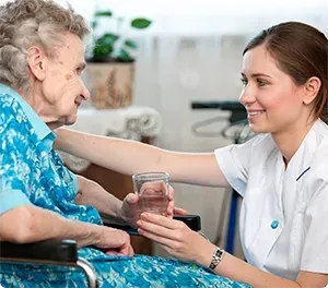 Woman in uniform offers water to elderly person in wheelchair, smiling. Interior setting.