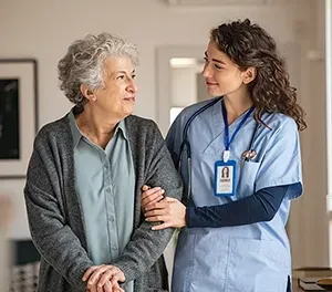 A caregiver assists an older person walking indoors. The caregiver wears scrubs and a stethoscope.