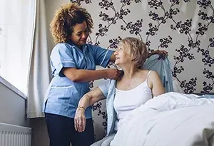 Caregiver assisting a person in a bed with putting on a robe. Bedroom setting.