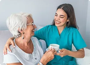 Caregiver offers a cup of tea to a smiling elderly person, both indoors.